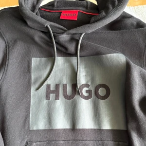 Svart hoodie från HUGO med logga - Svart hoodie från HUGO med stor grå logga framtill. Tröjan har huva med snörning, ribbade muddar och klassisk känguruficka. Tillverkad i mjuk bomullsmix som är perfekt för höst och vinter. Snygg och enkel design som passar till det mesta. Storlek xs men pass S då Hugo:S passform är lite större 