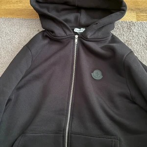 Svart hoodie från Moncler med dragkedja - Svart hoodie från Moncler med hel dragkedja framtill och klassisk huva. Tröjan har Moncler-logga på bröstet och är tillverkad i mjuk bomullsmix. Perfekt för en chill och stilren look.