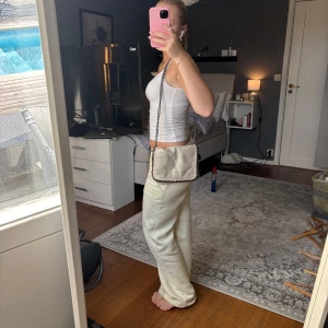 Beige axelväska med kedjerem - Säljer en snygg beige axelväska med cool kedjerem och dekorativa sömmar längs kanterna. Väskan har en mjuk, nästan glittrig yta och är perfekt för att lyfta vilken outfit som helst. Liten och smidig storlek, passar perfekt att bära crossbody. Finns inget märke men köpt på Öland 