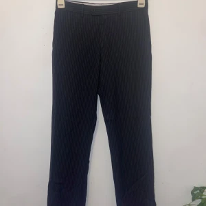 Randiga kostymbyxor från Zara Man - Snygga svarta kostymbyxor med tunna vita ränder från Zara Man. Byxorna har klassisk rak passform, bälteshällor och knäppning fram. Perfekta för en stilren look och passar till skjorta eller t-shirt. Materialet är mjukt och följsamt.