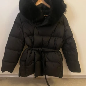 Svart pufferjacka äkta päls krage - Säljer en svart pufferjacka med stor fluffig äkta päls runt kragen och bälte i midjan. Jackan är vadderad och har en oversized look, perfekt för kalla dagar. Långärmad och riktigt mysig att bära under vintern. Jackan är använd ett fåtal gånger och är i ett bra skick.