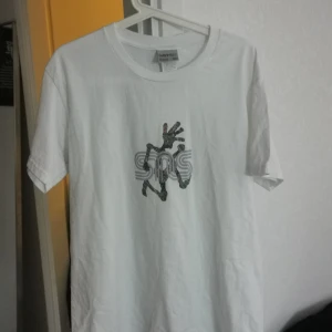 Vit t-shirt från Gramicci med tryck - Vit t-shirt från Gramicci med ett stort, färgglatt tryck på bröstet som föreställer en stiliserad figur och texten SNS. Klassisk passform och rund halsringning. Perfekt för dig som gillar streetwear och unika prints.