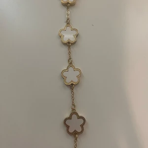 Guldarmband med vita blommor - Snyggt armband i guldton med flera vita blommor i klöverform. Blommorna har en fin guldkant med små detaljer som ger extra lyxig känsla. Perfekt accessoar för att lyfta din outfit med en söt och trendig touch.