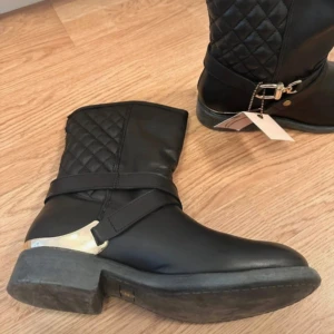 Svarta quiltade boots med gulddetaljer - Snygga svarta boots med quiltad detalj på skaftet och guldfärgade metalldetaljer vid hälen och spännet. Grov sula och låg klack ger en cool vibe. Perfekta för dig som vill sticka ut med stilrena men ändå unika skor