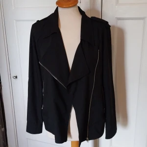 Svart bikerjacka från Xoul Edition XS - Svart bikerinspirerad jacka från Xoul Edition i storlek XS. Jackan har sned dragkedja, bred krage och axelklaffar. Långa ärmar med dragkedjor vid ärmsluten och två fickor framtill. Snygg och stilren modell i mjukt tyg.