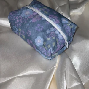 Blommig necessär i blått tyg - Handgjord Söt och praktisk necessär i blått tyg med blommigt mönster i lila, vitt och grönt. Väskan har en vit dragkedja upptill och en boxig form som rymmer mycket. Perfekt för smink, penslar eller småprylar när du är på språng.