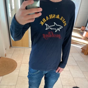 Mörkblå långärmad Paul & Shark tröja - Säljer en mörkblå långärmad tröja från Paul & Shark med tryck i gult, vitt och rött framtill. Tröjan har klassisk passform och är gjord i mjuk bomull. Perfekt för dig som gillar stilren design med sportig känsla och tydlig logga på bröstet.