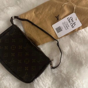 Louis Vuitton Monogram axelväska - Snygg axelväska från Louis Vuitton i klassiskt monogrammönster. Väskan är brun med LV-loggor och blommor, har dragkedja upptill och ett smalt axelband. Tillverkad i skinn och levereras med original dustbag. Perfekt för dig som vill ha en ikonisk och tidlös accessoar. Priset är lågt då kedjan skulle behöva bytas det kostar 300kr att göra. 
