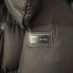 Svart lång dunjacka från Cedrico - Säljer en svart, lång och riktigt varm dunjacka från Cedrico. Jackan har en hög krage, dragkedja framtill och en snygg logga på bröstet. Perfekt för kalla dagar och har en klassisk pufferstil som är både stilren och praktisk.