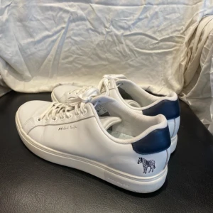 Paul Smith skor - Snygga vita sneakers från Paul Smith med mörkblå detalj på hälen och en liten zebralogga. Skorna har snörning och är tillverkade i skinn för en clean och stilren look. Perfekta för dig som gillar klassisk design med en twist.