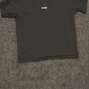 Svart BOSS t-shirt med logga - Svart t-shirt från BOSS med vit logga tryckt på bröstet. Klassisk rund hals och korta ärmar. Tillverkad i mjuk bomull för en skön känsla och enkel stil. Perfekt för dig som gillar minimalistiska plagg med tydlig branding.