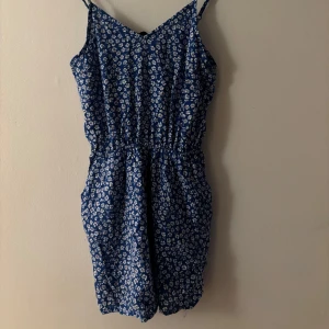 Blå blommig sommar byxdress H&M Divided - Supersöt blå kort byxdress från H&M Divided med vita små blommor, smala axelband och markerad midja. Perfekt för varma dagar och har en lätt och luftig känsla.  Byxdressen är ärmlös och har en avslappnad passform.