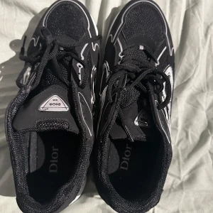 Svarta Dior B30 sneakers - Säljer ett par svarta Dior B30 sneakers med vita och silverfärgade detaljer. Skorna har tydlig CD-logga på sidan och bak, samt en chunky vit och svart sula. Ovandelen är i mesh och syntet med snörning framtill. Riktigt snygga och trendiga sneakers för dig som gillar lyxiga detaljer.