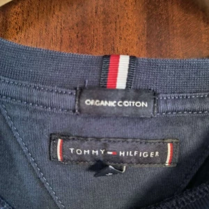 Blå långärmad tröja Tommy Hilfiger - Mörkblå långärmad tröja från Tommy Hilfiger i ekologisk bomull. Framsidan har det klassiska logotrycket med röd, vit och blå rand över bröstet. Rund halsringning och stilren design, perfekt för en clean och snygg look.