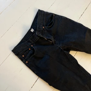 Svarta lågmidjade  bootcut jeans  - Snygga svarta jeans med bootcut-modell och klassisk femficksdesign. Jeansen har dragkedja och kopparfärgade nitar vid fickorna. Materialet är jeans och färgen är mörk svart, perfekt för en stilren look.