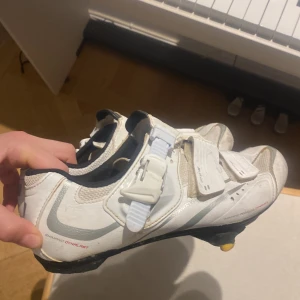 Vita Shimano cykelskor med spänne - Säljer ett par vita Shimano cykelskor med grå detaljer och svarta insidor. Skorna har kardborreband och ett kraftigt spänne för extra stabilitet. Utrustade med gula klossar för klickpedaler. Perfekta för dig som cyklar landsväg och vill ha grym kraftöverföring.