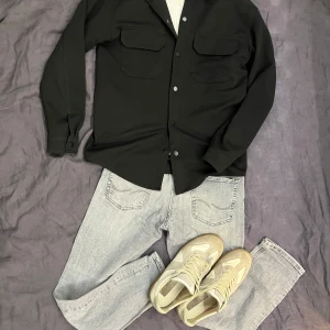 Svart overshirt  - Svart overshirt med klassisk krage, två stora bröstfickor med lock och knappar framtill. Skjortan har lång ärm och är tillverkad i ett mjukt bomullstyg. Perfekt att slänga över en t-shirt eller tröja för en chill look. Perfekt om du är runt 18-185