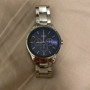 Silverfärgad klocka från Regal - Stilren klocka från Regal med blå urtavla och silverfärgat armband i metall. Länken är robust och har ett klassiskt spänne. Spännet har gått av men går att fixa på nån smyckes affär, och tror inte det kostar så mycket men där av priset. Skick 9/10 Inga repor eller liknande på glaset. Hör av dig vid minsta fundering och pris kan diskuteras vid snabb affär!