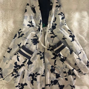 Blommig siphoodie från Benetton - Snygg siphoodie med blommigt mönster i marin och ljusblått på vit botten. Jackan har huva, dragkedja framtill, snörning och randiga muddar vid ärmsluten. Perfekt för dig som vill sticka ut med en cool och sportig look.