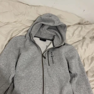 Grå hoodie från Sail Racing med dragkedja - Snygg grå hoodie från Sail Racing med hel dragkedja och extra bröstficka med dragkedja. Mjuk fleece på insidan och broderad logga på bröstet. Perfekt för kyliga dagar och riktigt skön att ha på sig.