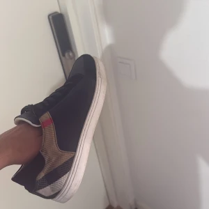 Burberry sneakers med rutigt mönster - Snygga svarta Burberry sneakers med klassiskt rutigt mönster i beige, svart och rött på hälen. Skorna har vit sula, svarta skosnören och rund tå. Perfekta för dig som vill ha en stilren men ändå unik look. Märkeslogga på plösen.