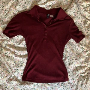 Vinröd ribbad polotopp med knappar - Vinröd polotopp från H&M med ribbad struktur och klassisk krage. Toppen har korta ärmar och knäppning framtill med fyra matchande knappar. Figurnära passform och mjukt material som känns skönt mot huden.