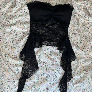 Svart spetsig korsett-topp - Säljer en svart korsett-topp med transparent spets och asymmetriska spetsdetaljer som hänger ner på sidorna. Toppen är axelbandslös och har en snygg bandeauform upptill med lätt rynkning i mitten. Toppen är vadderad och köpt i Grekland🌸Perfekt för dig som vill sticka ut med en edgy och elegant look✨