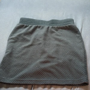 Svart kortkjol med struktur - En svart kortkjol med diskret strukturerat tyg och bred resår i midjan. Kjolen har en enkel och stilren design som är lätt att matcha med olika toppar. Perfekt för dig som gillar en klassisk look med en twist. Ettiket saknas