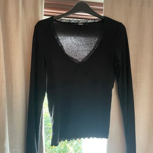 Svart långärmad topp med spets från Bik Bok - Svart långärmad topp från Bik Bok med v-ringning och spetsdetaljer längs halsringningen och under bysten. Modellen är ribbad och har en figurnära passform med vågig kant nertill. 