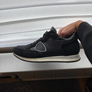 Philippe Model svarta sneakers - Svarta sneakers från Philippe Model med klassisk siluett. Skorna har ovandel i skinn och mocka, snörning och vit/grå sula. Sidan pryds av märkets ikoniska sköldlogga med vit kant. Perfekta för dig som gillar stilrena och sportiga skor.