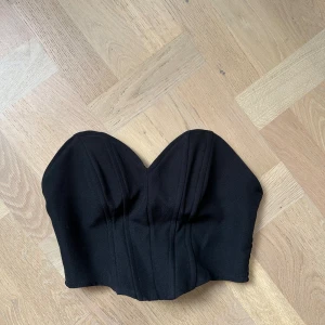 Svart korsett-topp från NA-KD - Säljer en svart korsett-topp från NA-KD med markerade sömmar och hjärtformad urringning. Toppen är axelbandslös och har en croppad passform som ger en snygg siluett. Perfekt att styla med höga jeans eller kjol.