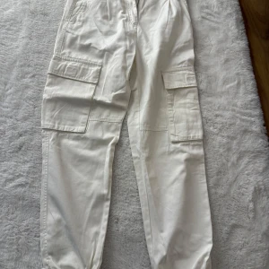 Vita cargobyxor från Bershka - Snygga vita cargobyxor från Bershka med elastisk midja och stora fickor på sidorna. Byxorna har en avslappnad passform och är perfekta för en trendig streetstyle-look. Tillverkade i mjuk bomull för extra komfort.