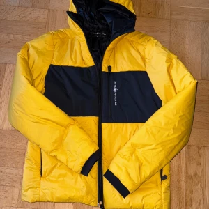 Gul och svart pufferjacka Sail Racing - Grym gul och svart pufferjacka från Sail Racing med huva och dragkedja. Jackan har en bred svart panel över bröstet och ryggen, samt svarta detaljer vid ärmslut och fickor. Tillverkad i syntetmaterial med Primaloft-foder som är både vindtätt och andas.