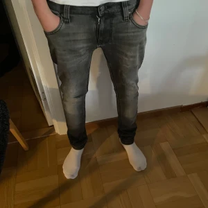 Grå jeans från Nudie Jeans - Säljer ett par grå jeans från Nudie Jeans med snygga slitningar och klassiska bakfickor med broderad detalj. Jeansen har normal midja och smal passform, perfekta för en trendig och avslappnad look. Modell tight terry