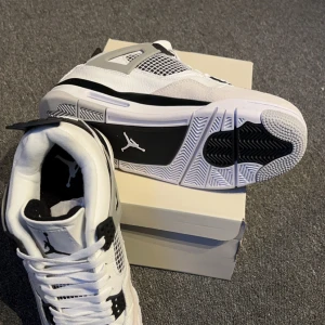 Nike Air Jordan 4 vit/svart - Säljer ett par Nike Air Jordan 4 i vit och svart med klassisk Jumpman-logga på plösen och hälen. Skorna har snörning, meshpaneler och detaljer i svart. Ovandelen är i skinn och mesh, med en platt sula och rund tå. Perfekta för dig som vill sticka ut med en ikonisk sneaker.