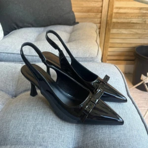 Svarta lackpumps med rosett - Snygga svarta pumps i lack med spetsig tå och elegant rosettdetalj framtill. Skorna har en smal klack och öppen häl med rem, vilket ger en stilren och trendig look. Perfekta för dig som vill sticka ut med en klassisk men modern stil.