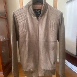 Beige Cardigan i mocka och stickat - Snygg beige Cardigan med framsida i mocka och ärmar samt rygg i stickad akryl. Jackan har ribbad krage, muddar och nederkant samt dragkedja framtill. Perfekt för dig som gillar en clean och stilren look. Storlek L. 