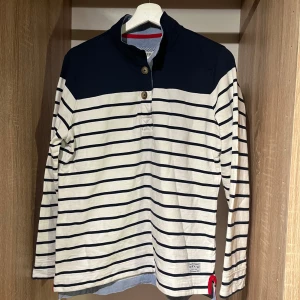 Randig pikétröja från Orvis, marinblå/vit - Snygg randig pikétröja från Orvis i marinblått och vitt. Tröjan har lång ärm, knappar vid halsen och hög krage. Sitter som en stor S eller liten M