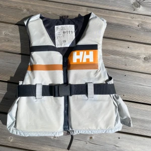 Helly Hansen flytväst  - Snygg flytväst från Helly Hansen i ljusgrått med svarta och orangea ränder. Västen har ett  justerbart midjebälte med spänne och dragkedja framtill. Knappt använd! Påse till flytvästen följer med😇Bild 4 visar en litet märke på ena bröstet.