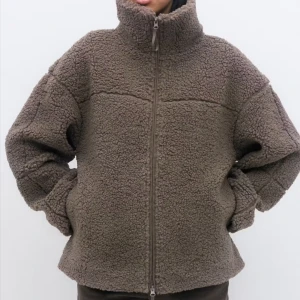 Brun teddyjacka med hög krage - Mysig brun teddyjacka med hög krage och hel dragkedja framtill. Jackan har en loose passform och är tillverkad i fluffigt fleece-material som håller dig varm. Perfekt för kyliga dagar och har långa ärmar samt diskreta sömdetaljer över axlar och rygg.