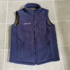 Mörkblå fleeceväst Dubbo Deer Norway - Mörkblå fleeceväst från Dubbo Deer Norway med broderad norsk flagga och logga på bröstet. Västen har hög krage, tre dragkedjeförsedda fickor och mjukt brunt teddyfoder på insidan. Perfekt för lager-på-lager under kyliga dagar.