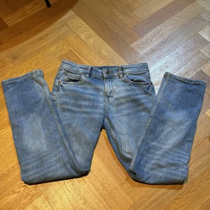 Blå jeans från Kappahl, storlek 152 - Klassiska blå jeans från Kappahl i storlek 152. Jeansen har fem fickor, bälteshällor och stängs med knapp och dragkedja. De är tillverkade i denim och har en rak passform med snyggt tvättad look. Perfekta för en avslappnad stil.
