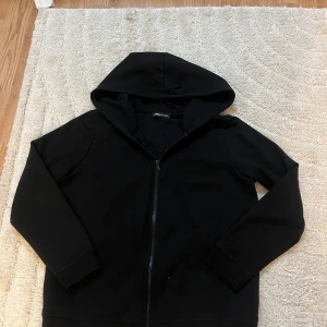 Svart hoodie med dragkedja Power Classic - Svart hoodie från Power Classic med hel dragkedja framtill och mysig huva. Tröjan har långärmade ärmar och är fodrad med mjukt material på insidan. Perfekt för kyliga dagar och enkel att matcha med allt.