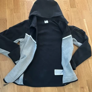 Nike hoodiejacka svart/grå XS - Nike hoodiejacka i svart och grått med dragkedja och huva. Jackan har en vit Nike-logga på bröstet, kontrasterande paneler och en ficka med dragkedja på ärmen. Tillverkad i mjuk bomullsblandning, perfekt för en sportig och avslappnad stil.