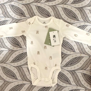 Vit långärmad body med nallemönster H&M - Supersöt vit långärmad body från H&M med små beige nallar som mönster. Tillverkad i mjuk bomull och har praktiska tryckknappar i grenen för enkel påklädning. Perfekt för mysiga dagar och passar till allt.