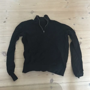 Svart tröja med half zip och struktur - Svart långärmad tröja med halv dragkedja i halsen. Framsidan har en strukturerad, stickad känsla medan ärmarna är släta. Perfekt för dig som gillar stilren och enkel design med en twist. Passar till både jeans och mjukisbyxor.