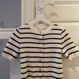 Randig kofta från H&M XS - Kortärmad kofta från H&M i storlek XS. Vit med svarta horisontella ränder och silvriga knappar framtill. Stickad i mjukt material med ribbad halsringning och ärmslut. Perfekt för lager-på-lager och en stilren look. Helt ny och aldrig använd. 
