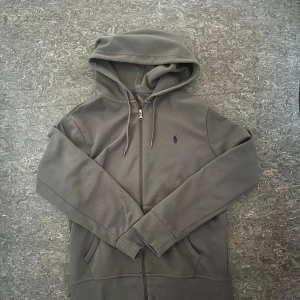 Grå Polo Ralph Lauren zip hoodie - Mörkgrå hoodie från Polo Ralph Lauren med dragkedja framtill, huva med snörning och klassisk broderad logga på bröstet. Tröjan har kängurufickor och är tillverkad i mjuk bomullsmix. Perfekt för chill dagar eller när du vill ha en clean streetstyle.