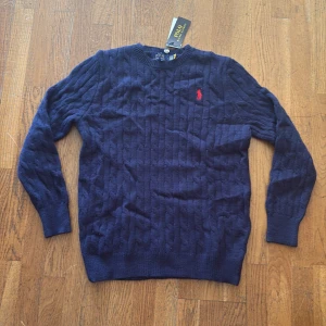 Mörkblå stickad tröja från Polo Ralph Lauren - Säljer en mörkblå stickad tröja från Polo Ralph Lauren med klassiskt kabelstickat mönster och röd broderad logga på bröstet. Tröjan har rund halsringning och långa ärmar, perfekt för lager-på-lager under kyliga dagar.