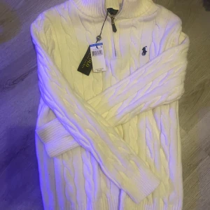 Vit kabelstickad tröja Polo Ralph Lauren - Kabelstickad vit tröja från Polo Ralph Lauren med halv dragkedja och hög krage. Klassisk design med broderad marinblå logga på bröstet. Mjuk och skön, perfekt för kyliga dagar. Långa ärmar och ribbade muddar för extra komfort.